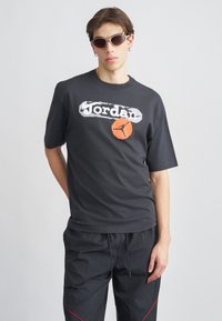 Μαύρο κοντομάνικο μπλουζάκι από βαμβάκι με λογότυπο "Jordan" σε λευκό και πορτοκαλί, μέσα σε ένα φθαρμένο γραφιστικό σχέδιο. Casual εφαρμογή.