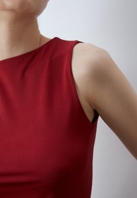 Top sin mangas rojo con una textura suave y un ligero brillo. Presenta un escote curvado, un diseño ajustado y un detalle asimétrico en el hombro.