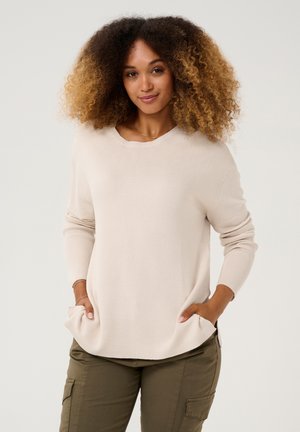 Beige ribbed svetr s dlouhými rukávy a bočními kapsami, s kulatým výstřihem a volným střihem, nošený přes olivově zelené cargo kalhoty.