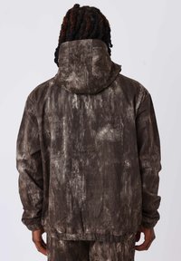 Giacca marrone con texture e cappuccio, caratterizzata da un motivo marmorizzato e polsini elasticizzati. Il tessuto sembra morbido e leggero.