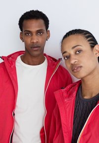 Chaquetas ligeras rojas con cremalleras blancas contrastantes, llevadas sobre una camiseta blanca y un suéter oscuro texturizado, exhibidas en dos modelos.