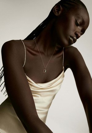 Collar de plata con un colgante único, lucido sobre un vestido de satén crema drapeado. El tono de piel es marrón oscuro, resaltando el contraste de la joyería.