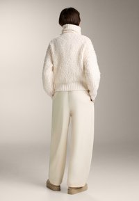 Krémový, štruktúrovaný pletený pulóver s vysokým golierom. V kombinácii s krémovobielymi širokými nohavicami a béžovými topánkami. Jednoduchá, uvoľnená silueta.