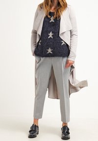 Pantaloni grigi a sigaretta, un pullover blu navy morbido con motivi di stelle argentate, e un giubbotto a strati grigio chiaro. Le scarpe nere con fibbia completano il look.