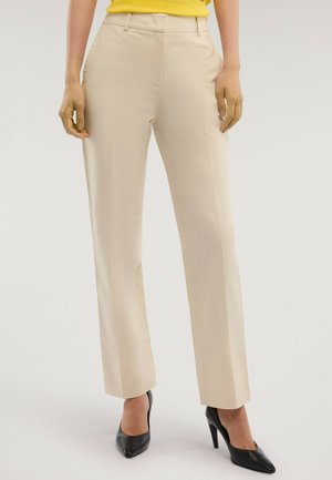 Kvinde iført beige bukser med lige ben, gul top og sorte højhælede sko, stående mod en ensartet lys baggrund.