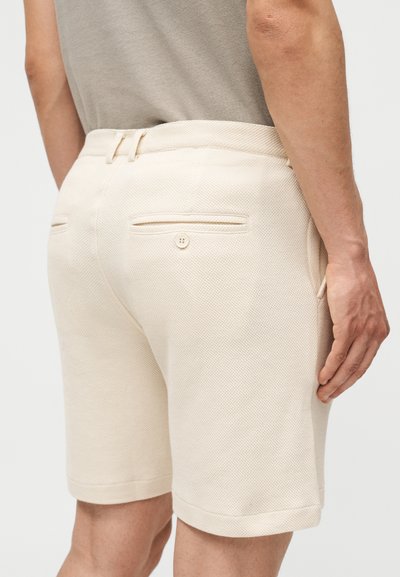 Μπεζ υφασμάτινες βερμούδες με κουμπωτή πίσω τσέπη, που φοριούνται από ένα άτομο με γκρι πουκάμισο, εμφανιζόμενο από πίσω στο ύψος των μηρών.