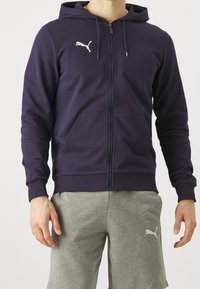 Námořnická zipová mikina s klokánkovými kapsami a žebrovanými manžetami. Má bílé logo Puma na levé části hrudníku. Kombinováno se šedými šortkami.