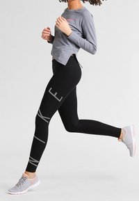 Maglietta grigia a maniche lunghe e leggings neri con strisce reflective, abbinati a scarpe da ginnastica grigie. I leggings hanno un design aderente e un materiale elasticizzato.