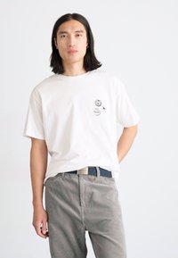 T-shirt bianco in cotone con scollatura rotonda, vestibilità ampia, con tre toppe grafiche sul lato sinistro del petto. Abbinato a pantaloni in velluto a coste grigi.