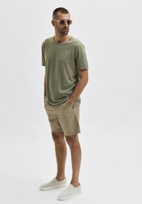 Olivgrön randig t-shirt, beige shorts med fickor och ljusa slip-on skor. Tillbehören inkluderar solglasögon med genomskinlig ram.