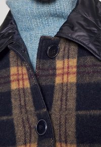 Ullblandad jacka i mörkblått med ett rutmönster i gult och rött. Har en krage i svart nylon och knappstängning. Ljusblå stickad tröja under.