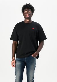 T-shirt en coton noir avec un logo rouge sur la poitrine gauche, associé à un jean bleu usé. La coupe est décontractée avec des manches courtes.