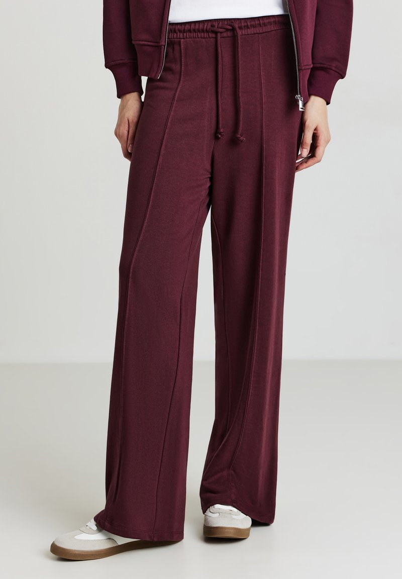 Pantaloni larghi bordeaux con vita elastica, dettagli cuciti e cordoncino; abbinati a sneakers bianche. Tessuto morbido e vestibilità rilassata.