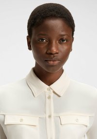 Light cream button-up shirt with a collar, featuring two front pockets and a smooth texture. The fabric appears soft and lightweight.