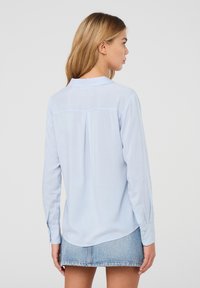 Camicetta azzurro chiaro a maniche lunghe con colletto a punta, orlo dritto e piega singola sul retro, realizzata in un tessuto leggero e liscio.