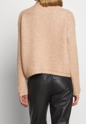 Pull en grosse maille beige porté par une personne aux cheveux courts, associé à un pantalon en cuir noir, vu de dos sur fond uni.