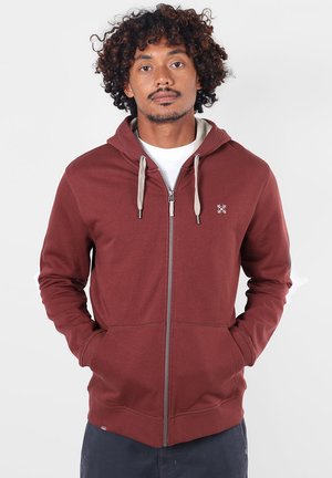 Homme aux cheveux bouclés portant un sweat à capuche bordeaux zippé avec des cordons beiges et les mains dans les poches, sur un fond uni.