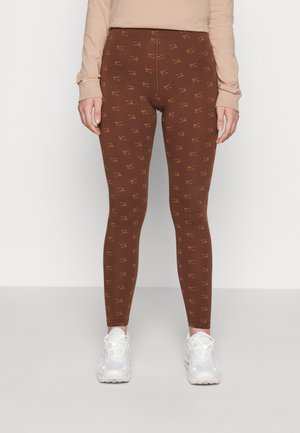 Person iført brune leggings med gentagne gule Nike Air-logoer og hvide sneakers mod en ensfarvet hvid baggrund.