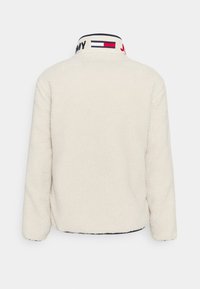 Giacca in fleece beige chiaro con colletto alto. Presenta un colletto brandizzato in blu navy, rosso e bianco. Texture liscia e vestibilità comoda.