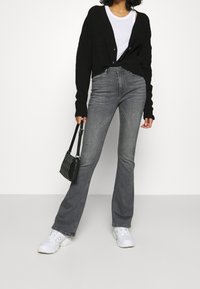 Persona che indossa un cardigan nero, una camicia bianca, jeans bootcut grigi e sneakers bianche, mentre tiene una borsa nera su uno sfondo neutro.