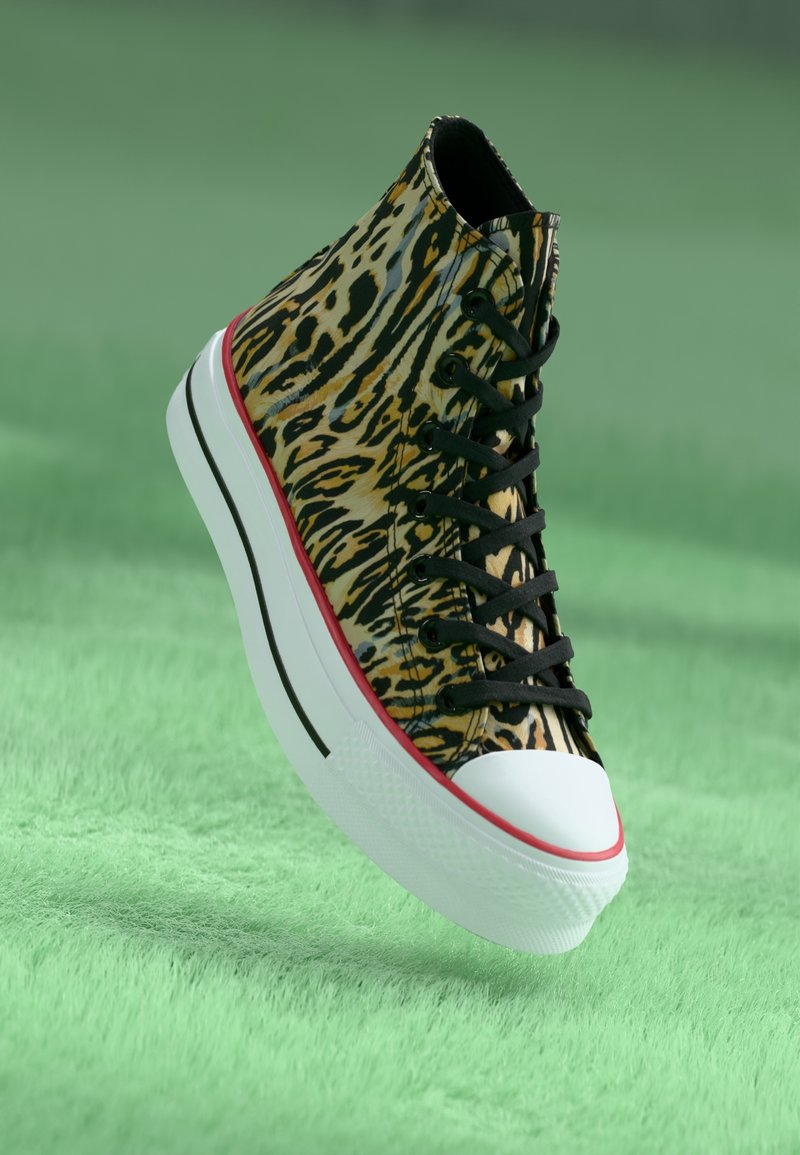 Sneaker montante avec tige imprimée léopard, lacets noirs, bout blanc et semelle blanche épaisse avec bordure rouge et noire sur surface texturée verte.