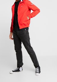 Giacca rossa con zip e colletto blu navy e bianco, indossata sopra una maglietta nera; abbinata a jeans neri e sneaker alte nere.