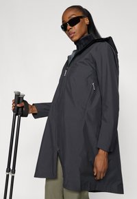 Vandafvisende sort jakke med lynlås foran, hætte og sidelommer. Modellen holder på gåstave og har et strømlinet design samt en glat tekstur.