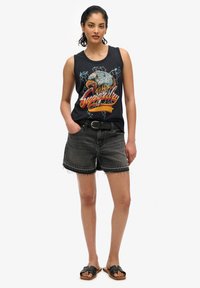 Canotta nera in cotone con grafica di un'aquila, pantaloni corti in denim grigio con orlo sfrangiato e sandali neri con fasce larghe.