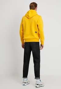 Man som bär en ljusgul hoodie, svarta uppvikta jeans, vita sneakers med ljusblå detaljer, står vänd bort från kameran mot en enkel bakgrund.