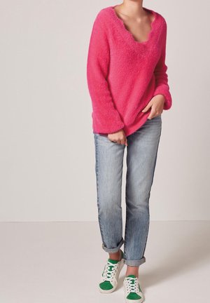Persona que lleva un suéter peludo de color rosa brillante, vaqueros azules claros con puños y zapatillas blancas con detalles verdes sobre un fondo liso.