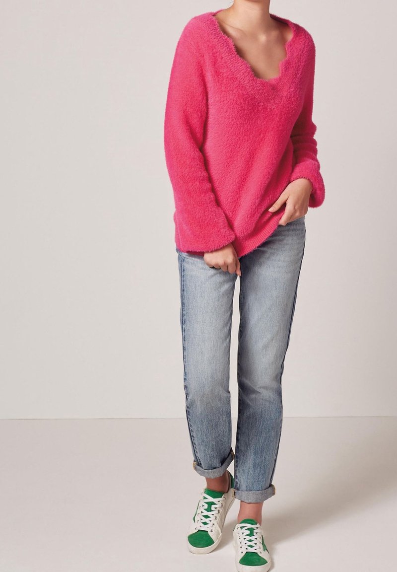 Personne portant un pull rose vif en laine duveteuse, un jean bleu clair retroussé et des baskets blanches avec des accents verts sur un fond uni.