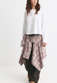 Weißes, verkürztes Langarmshirt, kombiniert mit einem rosa karierten Flanellhemd, das um die Taille gebunden ist, schwarzen Hosen und braunen Schuhen, ergänzt durch einen gemusterten Gürtel.