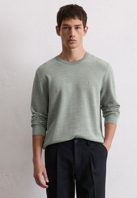Pull en maille vert clair avec un col rond et des manches longues, associé à un pantalon noir ajusté. Un motif texturé subtil est visible.