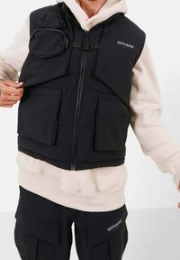 Schwarze gesteppte Weste mit Frontreißverschluss, ausgestattet mit zwei Taschenklappen und mehreren Vordertaschen. Wird über einem beigen Hoodie und passenden Hosen getragen.