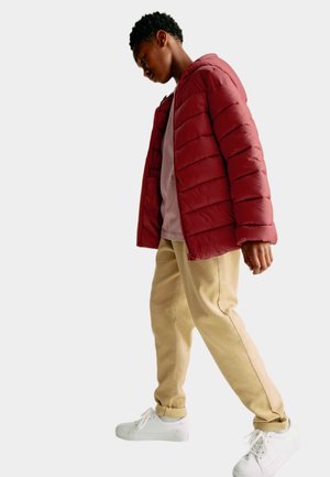 Ung person iført en rød puffjakke, beige bukser og hvide sneakers, der går med hovedet lidt nedad foran en ensfarvet baggrund.
