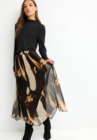 Kvinde med langt hår, iført en sort bluse med lange ærmer og en plisseret, flydende midi-nederdel i sort og beige mønster, mens hun holder i nederdelens side.
