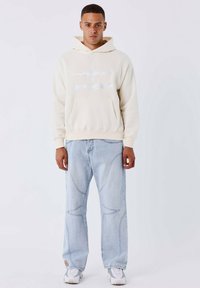 Crème hoodie met een voorzak en logo, gedragen met lichtblauwe wide-leg jeans met contrasterende naaddetails en witte sneakers.