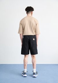 Beige T-Shirt mit kurzen Ärmeln kombiniert mit schwarzen Cargo-Shorts. Sichtbares Markenlogo auf den Shorts. Weiße Socken und Sneakers mit blauen Akzenten.