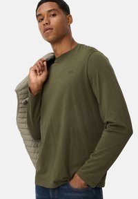 Olijfgroene longsleeve shirt met een ronde hals en subtiele logo-detail, gecombineerd met een gestructureerde lichtgrijze jas die van de schouder hangt.