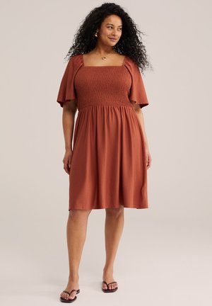 Femeie cu păr creț purtând o rochie căprui spre ruginiu, până la genunchi, cu mâneci evazate și sandale negre, stând în fața unui fundal simplu.