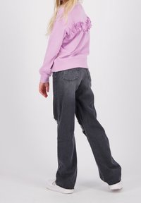 Lila sweatshirt med fransdetalj på baksidan, tillsammans med höga, löst sittande mörkgrå jeans och vita sneakers.