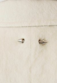 Tessuto di lana bianca con due borchie metalliche argento a forma di punta. Texture morbida con un aspetto leggermente peloso e dettagli cuciti.