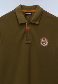 Polo vert olive avec deux boutons, détail orange près du col et logo rond brodé sur la poitrine gauche.
