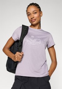 Light purple t-shirt with a round graphic design in white, short sleeves, and a relaxed fit. Worn with a black backpack.