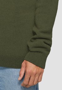 Gebreide groene sweater met ribbels aan de manchetten en de onderkant. Het materiaal lijkt zacht met een gestructureerde afwerking. Een hand ligt op de spijkerbroek.