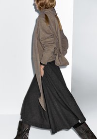 Brauner Strickpullover und langer schwarzer karierten Rock, kombiniert mit dunkelbraunen kniehohen Stiefeln. Das Outfit besticht durch strukturierte Stoffe und ein mehrlagiges Design.