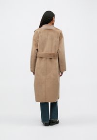 Ilgas smėlinis paltas su švelnia tekstūra, plačiais atlapais, dirželiu talijoje ir nukritusiais pečiais, derinamas su tamsiai mėlynomis plačiomis džinsomis ir juodais batais.