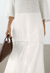 Maglione bianco a maniche corte, abbinato a una gonna bianca fluida e a una borsa a tracolla intrecciata marrone, che mette in risalto la texture e il design a strati.