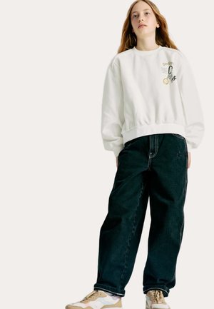 Sweatshirt blanc avec ceinture élastique et graphiques brodés, associé à un jean ample foncé et des baskets multicolores.