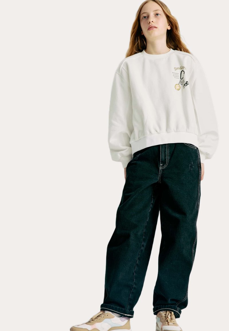 Sweatshirt blanc avec ceinture élastique et graphiques brodés, associé à un jean ample foncé et des baskets multicolores.